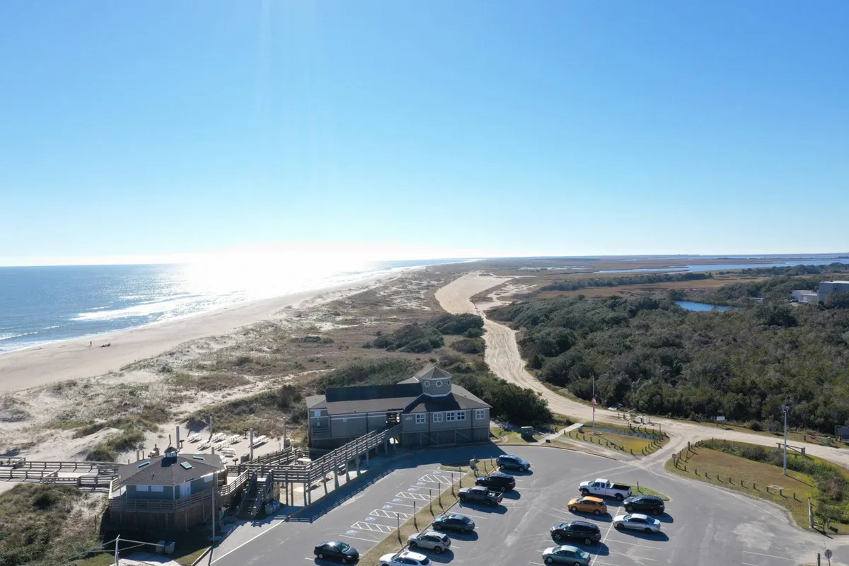 A wide exterior view of Carolina Beach shows the boardwalk, sandy beach, ocean, and ample parking area. The image conveys the ease of accessing beachside activities, walking to the boardwalk, and using the parking space for vehicles or golf carts. The dunes and ocean in the background emphasize the quintessential coastal experience.