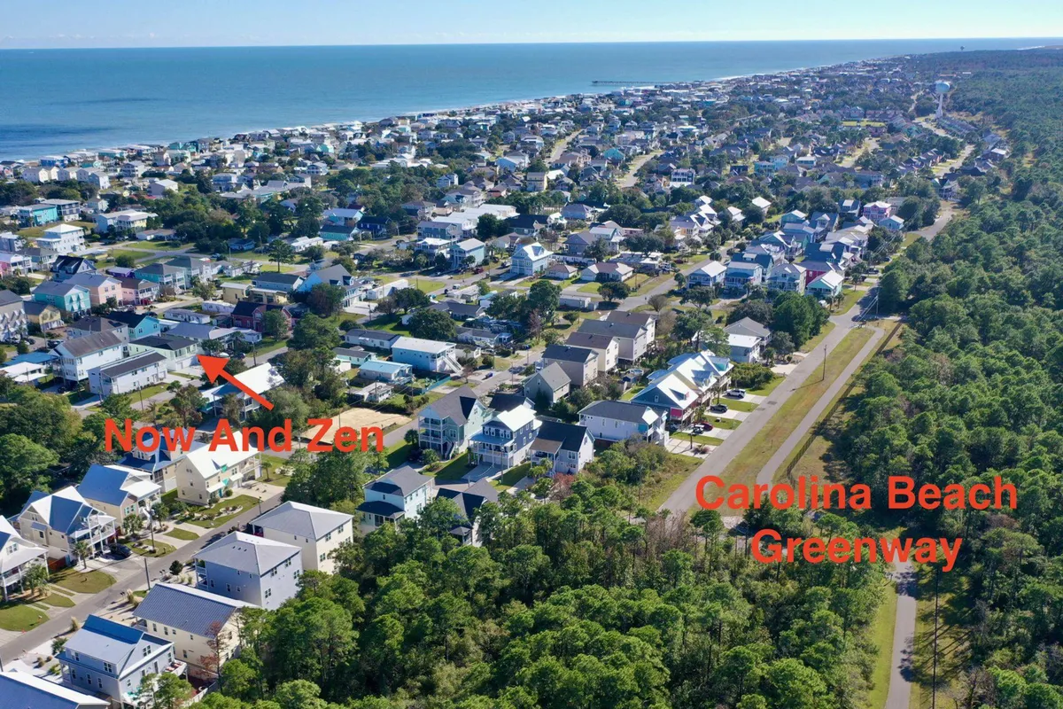 The exterior view highlights the Now And Zen property situated next to the Carolina Beach Greenway, with the ocean visible in the distance and a tranquil residential area in the foreground. The greenway offers ample space for walking or biking, ideal for dog owners seeking scenic strolls with their pets.