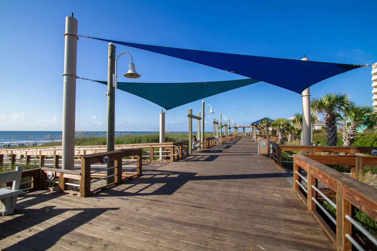 A scenic shot of a wooden boardwalk lined with shade sails and palm trees, all leading towards a sparkling ocean view. The setting feels relaxed yet social, hinting at opportunities for strolls, gatherings, or simply enjoying a beautiful sunset by the sea.