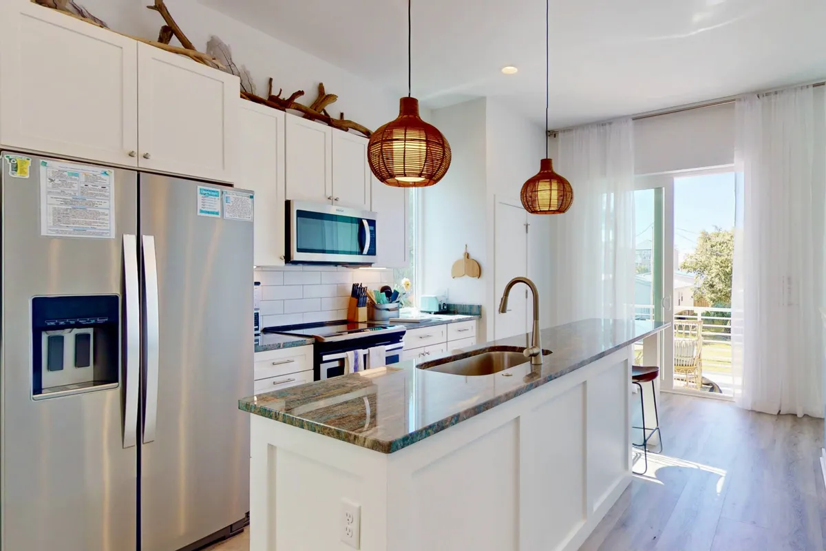 A contemporary kitchen with a spacious island countertop, stainless steel appliances, and elegant hanging pendant lights. The sliding glass door offers a glimpse of the outdoors, creating a sense of flow and openness. The space is set up for cooking, entertaining, and family gatherings, filled with natural morning light.