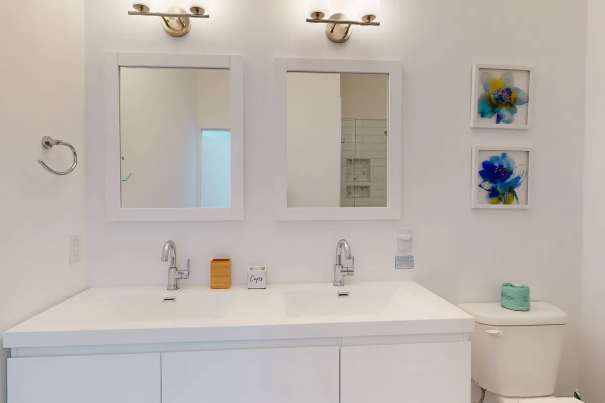 A modern, calming bathroom with dual sinks, large mirrors, soft artwork, and stylish lighting. The space is organized and bright, offering plenty of room for morning routines or evening wind-downs.