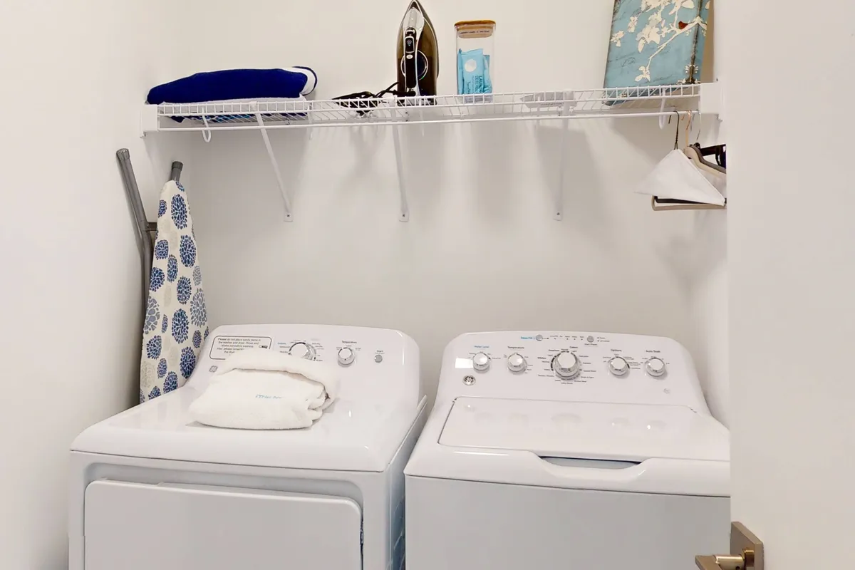 A neat laundry nook with a modern washer and dryer set, an iron placed nearby, and a decorative shelf above. The clean lines and uncluttered space highlight household practicality, while small touches like decor on the shelf add a sense of home.