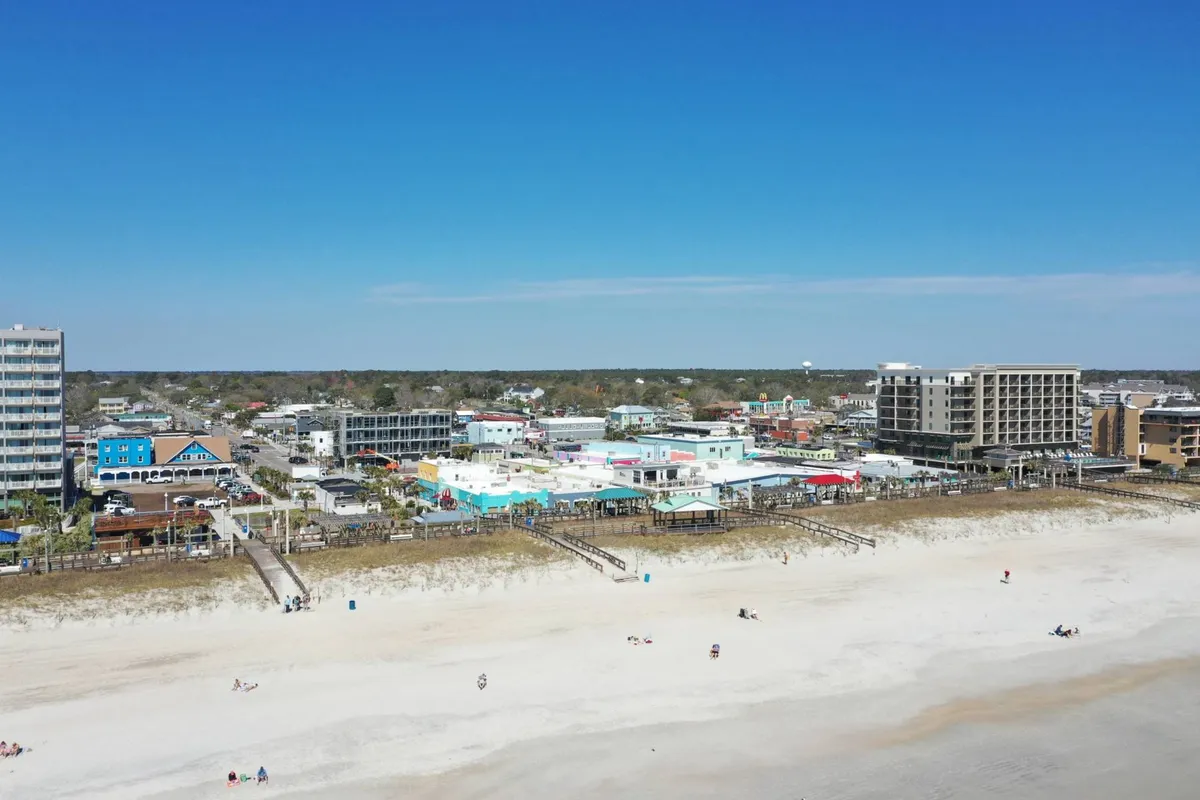 This image captures the vibrant Carolina Beach Boardwalk area, with the beach, colorful buildings, and people enjoying various activities. The lively scene showcases families relaxing, exploring shops, and participating in beachside fun, all under a sunny sky.