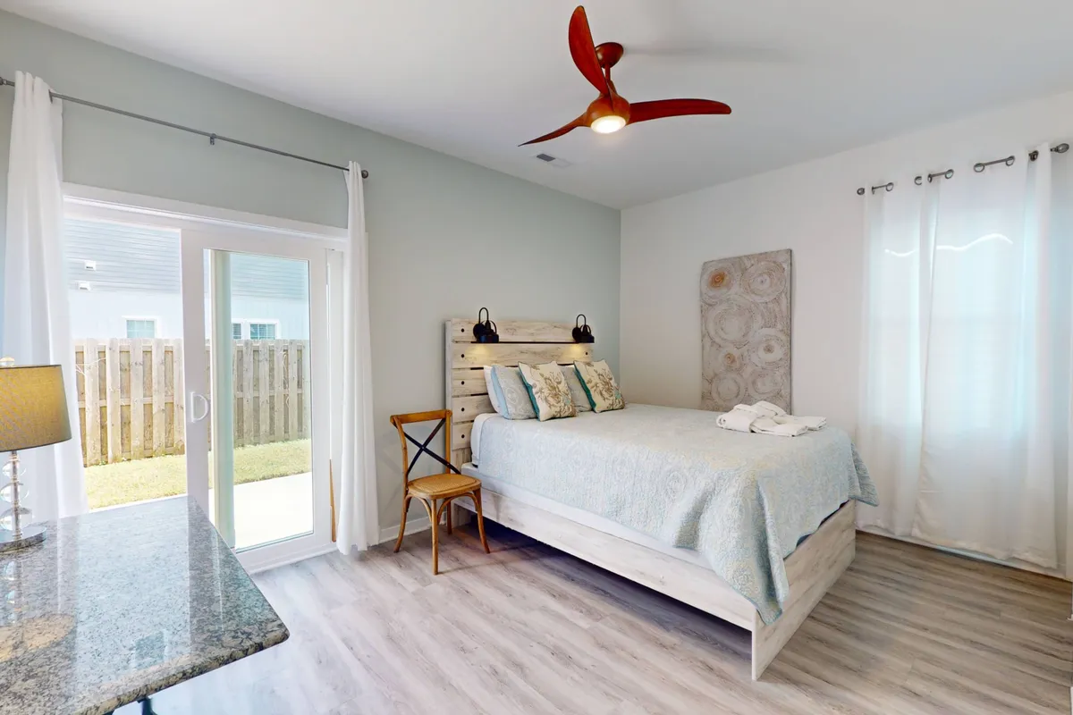 A cozy queen bedroom showcasing a wooden headboard, decorative pillows, a modern ceiling fan overhead, and a large window with sheer curtains allowing natural light to filter in. The space feels both inviting and functional, with touches designed for comfort.