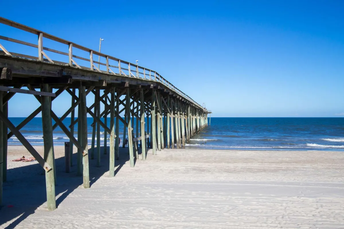 A picturesque view of the pier extending over the ocean, with soft sand and clear skies. People can enjoy walking, fishing, or relaxing by the water, representing quintessential local activities that groups and friends can enjoy together.