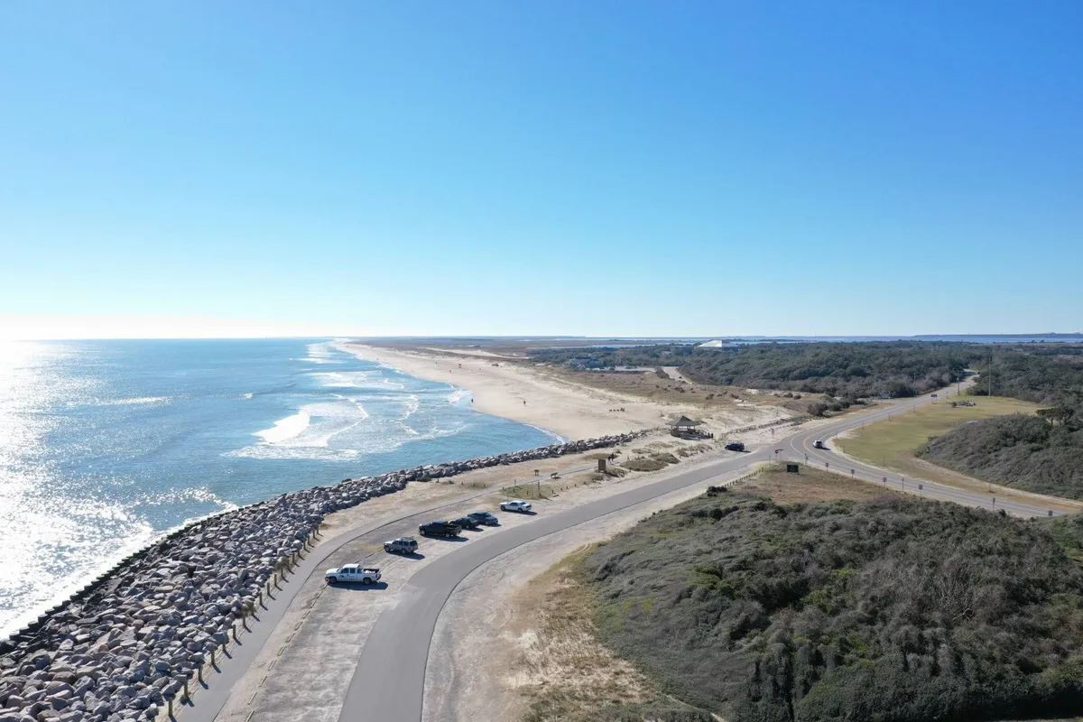 A sweeping exterior view of the sandy beach with gentle ocean waves, parked vehicles nearby, and a jetty extending into the water. The scene evokes a sense of openness and tranquility, with surfers catching waves and seashells scattered along the shore under a serene sky.