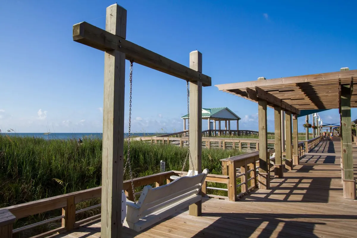 A peaceful exterior setting featuring a wooden porch swing under an overhead structure, overlooking a boardwalk and a sweeping view of the ocean. The scene is bathed in soft light, inviting guests to relax, listen to the waves, and soak up the tranquil atmosphere.