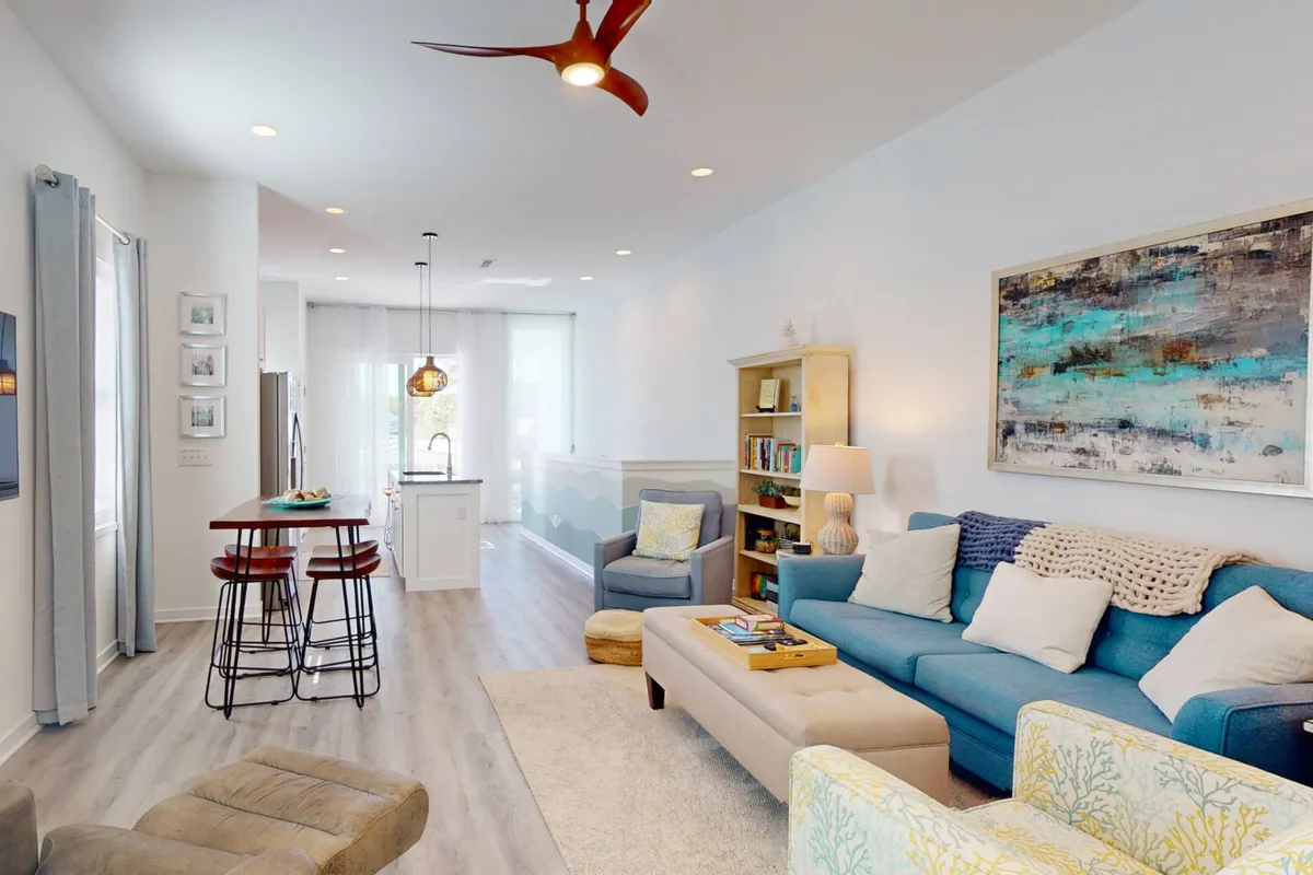 The image showcases a bright, inviting living space featuring a plush blue sofa, a stylish wooden bookshelf, a sunlit kitchen island with sleek bar stools, and vibrant coastal-inspired artwork. The décor blends beachy tones of blue and white with high-end finishes, creating a soothing yet sophisticated atmosphere—perfect for relaxation or gathering with friends and family.