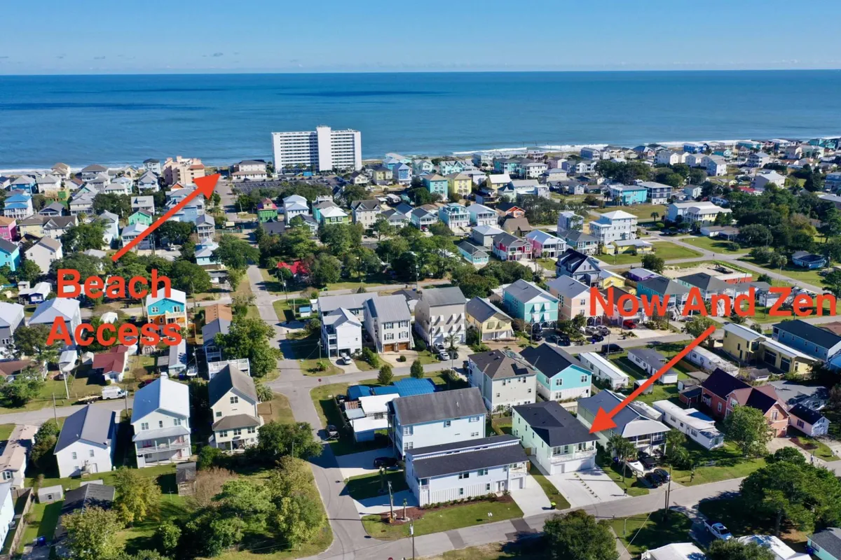 The exterior of the Now And Zen property is visible, nestled among other colorful beach houses with the ocean in view. The scene shows easy access to the beach and hints at outdoor gathering spaces perfect for group relaxation or barbecues. The atmosphere is vibrant yet tranquil, with the home positioned just steps from the sand.