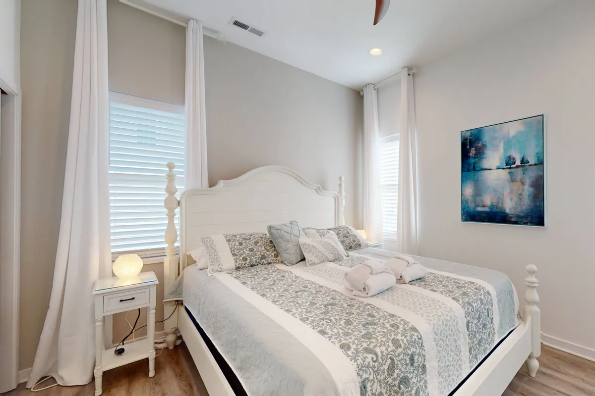A serene bedroom featuring a neatly made bed with decorative pillows, soft artwork above the headboard, a nightstand with a lamp, and subtle, calming décor. The overall atmosphere is tranquil and inviting, ideal for rest and relaxation.