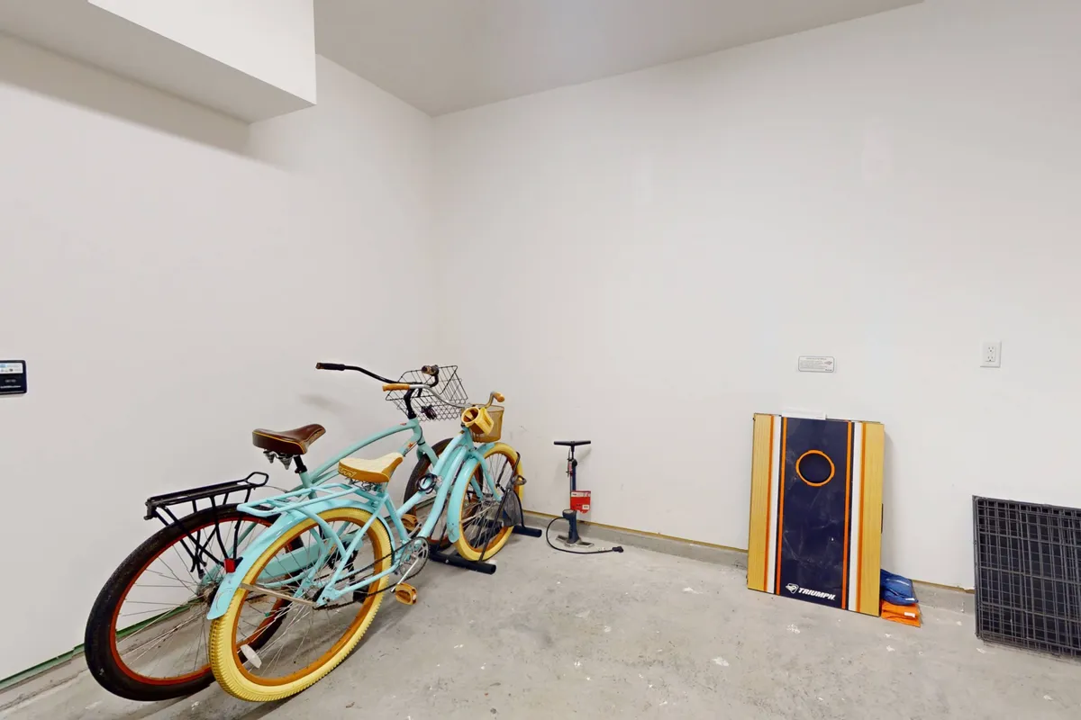 A bright, organized garage space featuring two bicycles propped against the wall, a bicycle pump, and a cornhole board ready for play. The floor is tidy, and there’s ample room for storage, with the cornhole board hinting at recreational opportunities. The garage conveys both practicality and leisure.