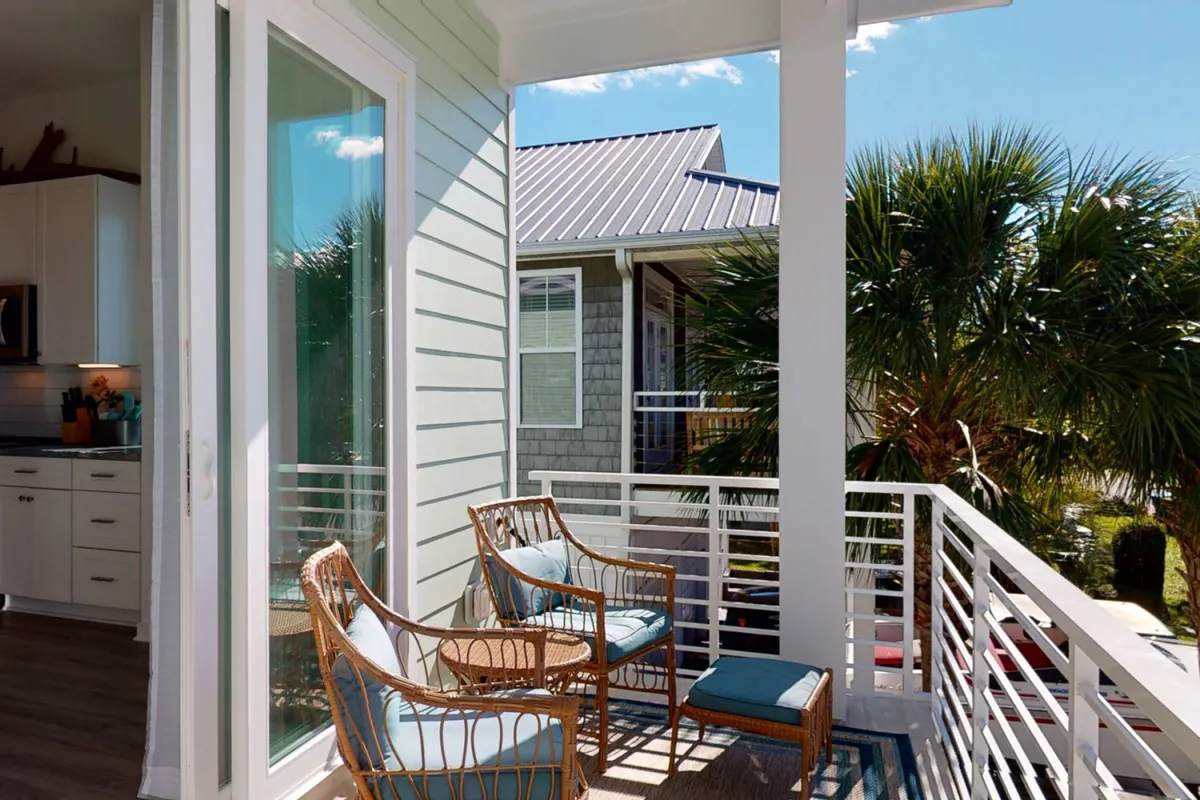 An exterior scene featuring a calm balcony with wicker chairs and a palm tree, with a neighboring property in the distance. The setting is cozy and ideal for morning coffee or evening cocktails, echoing the laid-back beach lifestyle.